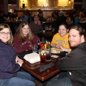 Four people smiling at a table in a busy restaurant, enjoying a trivia night with meals in front of them.