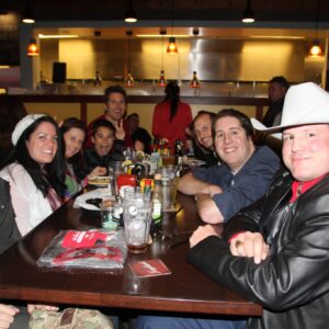 A group of people smiling around a dinner table in a restaurant, with one man wearing a white cowboy hat. Glasses and beverages are visible on the table as they enjoy a DJ trivia night.