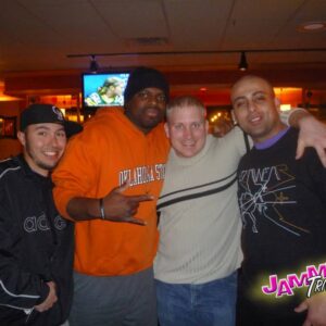 Four men smiling and posing together at a bar with a television in the background displaying a sports game.