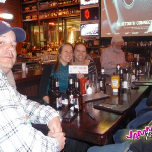 Group of people sitting at a bar with drinks, participating in pub trivia, smiling at the camera; bar signage and TV screens are visible in the background.