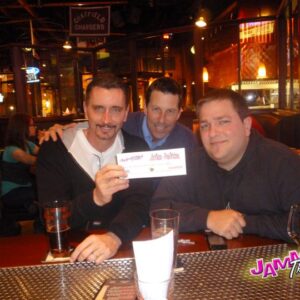 Three men smiling at a table in a bar, holding a DJ trivia night prize certificate, with the venue bustling in the background.