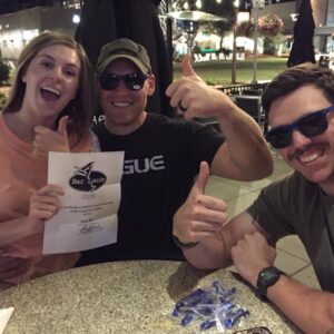 Three friends smiling at a table outside, one holding a trivia paper, with thumbs up, at night under lights.