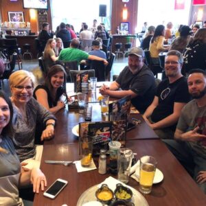 A group of six adults, smiling and seated around a table in a busy restaurant, participating in DJ trivia, with drinks and menus visible.