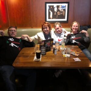 Four adults in matching "moron" t-shirts pose happily around a pub table with drinks, food, and a photo of a couple on the wall behind them during a DJ trivia night.