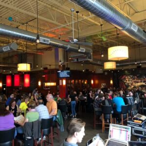 A busy restaurant interior with patrons enjoying pub trivia, colorful hanging lights, a bar on the right, and industrial ceiling elements.
