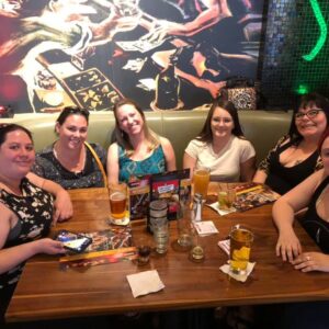 Six women smiling at a table in a bar with drinks and menus, a colorful mural on the wall behind them, enjoying a DJ trivia night hosted by a popular trivia company.