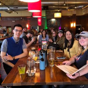 Group of happy people sitting at a bar, smiling at the camera, with drinks and menus on the table in a warmly lit setting, enjoying a lively round of pub trivia.