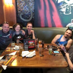 Five friends enjoying a meal together at a restaurant booth, smiling and posing for a photo during pub trivia, with drinks and menus on the table.