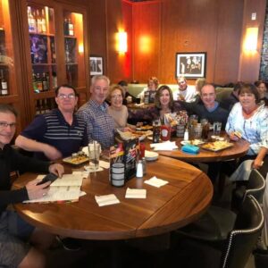 Seven people smiling around a restaurant table filled with food, in a warmly lit room with artworks on the walls during a DJ trivia night.