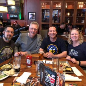 Four adults smiling at a table in a bustling pub during trivia night, with food and drinks visible.