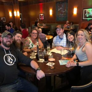 Group of six adults smiling at a table in a restaurant with drinks, in a lively atmosphere with trivia on the TV in the background.