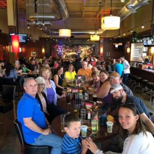 A group of people of various ages smiling at a table in a busy pub trivia event with warm lighting and contemporary decor.