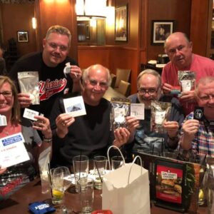 Group of six adults celebrating with drinks and showing off playing cards and other small items at a table during pub trivia in a restaurant.