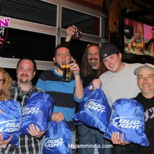 Group of seven adults smiling and posing at a pub trivia night event in a bar, holding Bud Light merchandise and beer bottles, with event logos displayed.