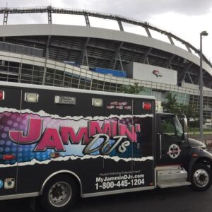 A dj trivia service van branded "jammin' djs" parked outside a large stadium on an overcast day.
