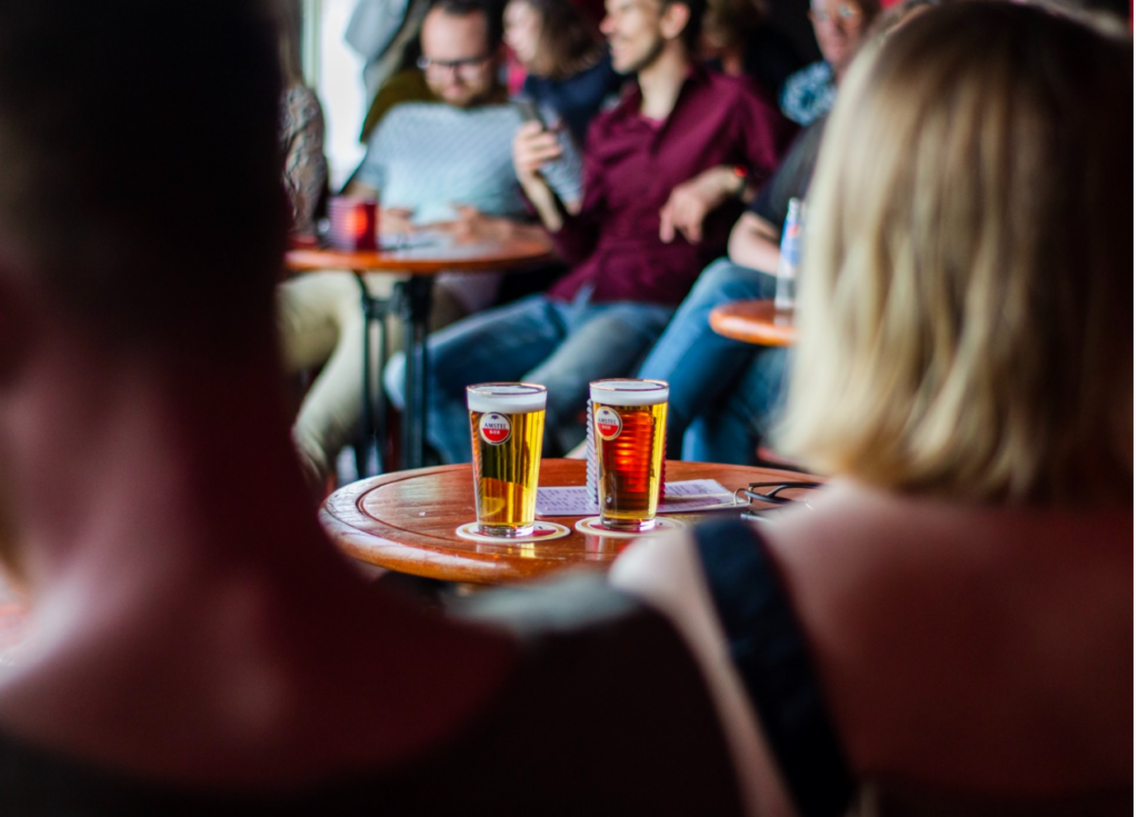 Two glasses of beer on a table in a bustling pub, with people engaged in pub trivia in the out-of-focus background.