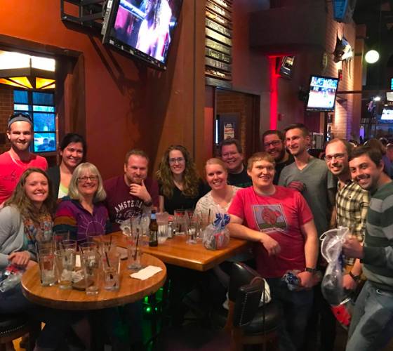 Group of friends smiling at a table in a bar with drinks, casually dressed, playing dj trivia with TVs and orange walls in the background.