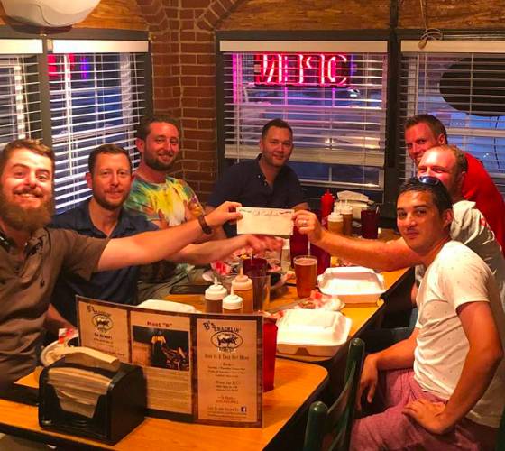 Group of seven men smiling and toasting at a pub trivia night, illuminated by warm light with neon signs visible in the background.