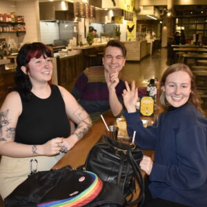 Three friends smiling and sitting at a table in a food court, one gesturing a peace sign.