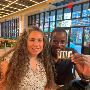 A man and a woman sitting at a table indoors, smiling at the camera; the man is holding up a card that says "trivia.