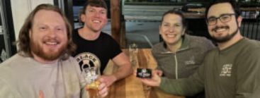 Four men smiling at a pub trivia event, three holding beer glasses, with a window in the background.