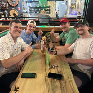 Four men smiling and sitting around a wooden table in a bar, giving a thumbs up, with drinks and snacks on the table during a dj trivia night.