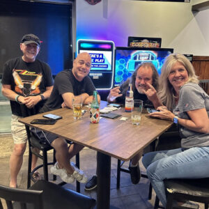 Four adults smiling at a table in a sports bar with drinks, sports on TV in the background, and pub trivia visible.