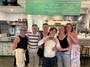 A group of six people stand smiling inside a restaurant. A young boy at the center holds a black wallet, and a menu board is visible in the background.