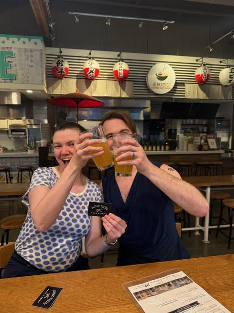 Two people are smiling and clinking glasses of beer at a wooden table in a modern bar with Asian decor. One holds a sign that reads "JUNCTION.