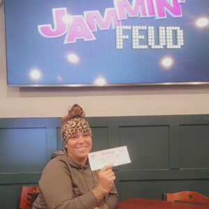 A woman sitting at a table holds up a check and smiles in front of a screen displaying the words "Jammin' Feud.