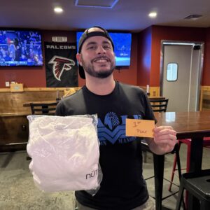 A man smiles while holding a white packaged item in one hand and a card reading "1st Place" in the other, standing in a bar with sports decor and TV screens in the background.